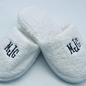 May include: A pair of white terry cloth slippers with the embroidered initials "MJG" in dark blue thread. The slippers are fluffy and appear soft, suggesting comfort and relaxation. The background is plain white.