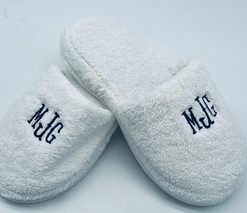 May include: A pair of white terry cloth slippers with the embroidered initials "MJG" in dark blue thread. The slippers are fluffy and appear soft, suggesting comfort and relaxation. The background is plain white.