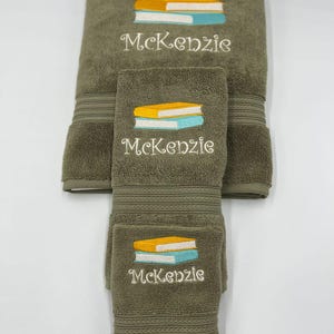 May include: A set of three green towels with embroidered stacks of books and the name "Kenzie".