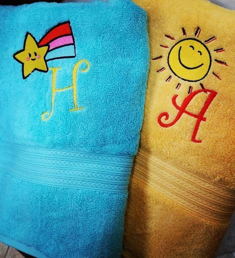 May include: Two embroidered towels, one blue with a yellow star and rainbow, the letter "H" and the other yellow with a yellow sun and the letter "A".