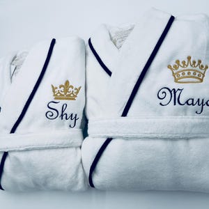 May include: Two white bathrobes with black trim and gold embroidered crowns. One robe has the name "Shy" embroidered on it, and the other has the name "Maya" embroidered on it.