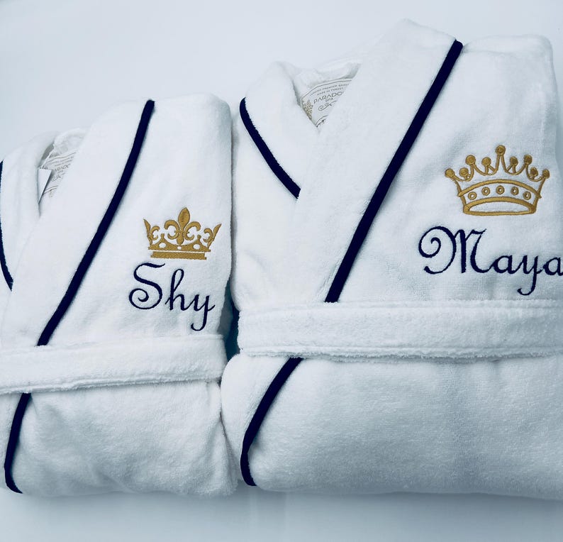 May include: Two white bathrobes with black trim and gold embroidered crowns. One robe has the name "Shy" embroidered on it, and the other has the name "Maya" embroidered on it.