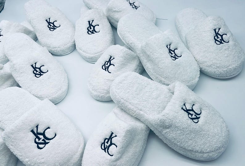 May include: A collection of white terry cloth slippers, each embroidered with a black monogram of the letters "K&C". The slippers are arranged on a light-colored surface, showcasing their soft texture and design.