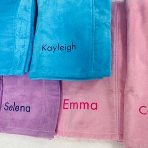 May include: A collection of personalized plush blankets in various pastel colors, including blue, purple, and pink. Each blanket is embroidered with a name in a contrasting color. The names include Baylie Grace, Kayleigh, Selena, Emma, and Caroline.