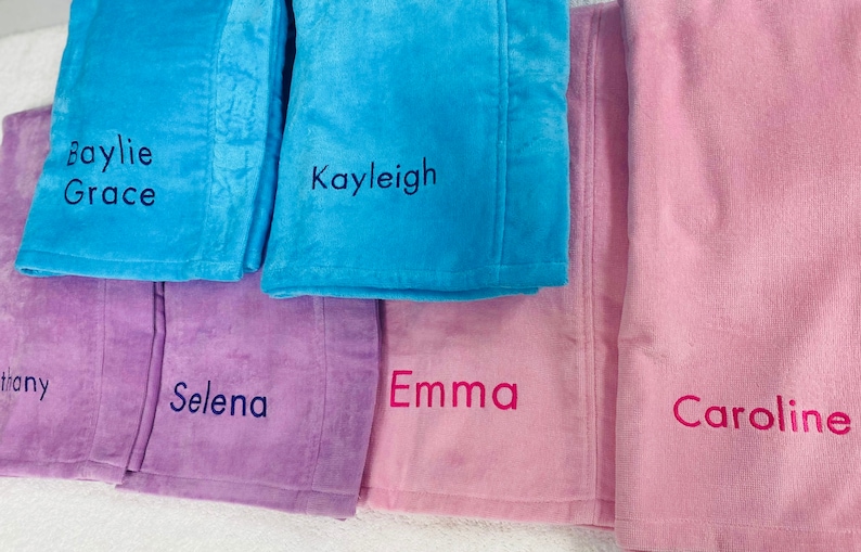 May include: A collection of personalized plush blankets in various pastel colors, including blue, purple, and pink. Each blanket is embroidered with a name in a contrasting color. The names include Baylie Grace, Kayleigh, Selena, Emma, and Caroline.