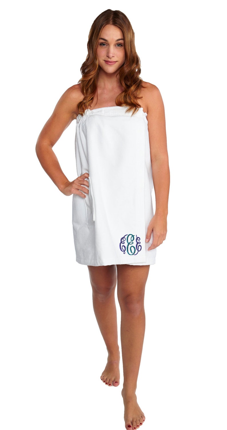 May include: White spa wrap with a pocket and an embroidered monogram in blue and teal. The wrap is knee-length and appears to be made of soft, absorbent material. The model is wearing the wrap.