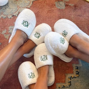 May include: White terry cloth slippers with embroidered initials in green thread. The slippers are shown on multiple feet, with the initials "MSK", "SME", and "KPD" visible. The slippers are on a patterned rug.