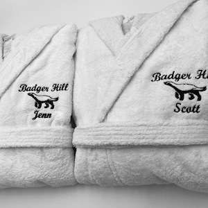 May include: Two white bathrobes with black embroidered text and an image of an animal. The text reads "Badger Hill" and the names "Jenn" and "Scott".