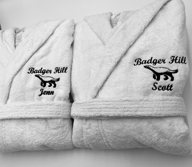 May include: Two white bathrobes with black embroidered text and an image of an animal. The text reads "Badger Hill" and the names "Jenn" and "Scott".