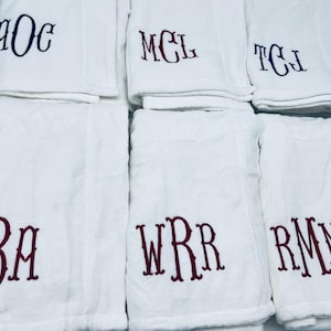 May include: White bath towels with embroidered initials in various colors. The towels are folded and stacked, with the initials "AOC", "MCL", "TCJ", "LBA", "WRR", and "RMN" visible. The towels are made of a soft, absorbent material.