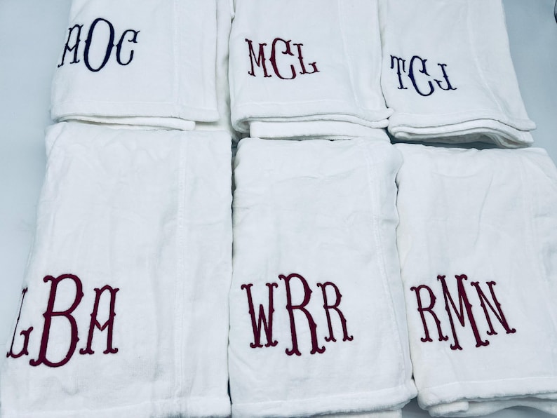 May include: White bath towels with embroidered initials in various colors. The towels are folded and stacked, with the initials "AOC", "MCL", "TCJ", "LBA", "WRR", and "RMN" visible. The towels are made of a soft, absorbent material.