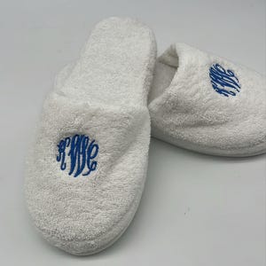 May include: A pair of white terry cloth slippers with a blue embroidered monogram. The slippers are fluffy and soft, perfect for lounging. The monogram is a stylized script design.