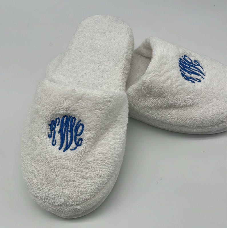 May include: A pair of white terry cloth slippers with a blue embroidered monogram. The slippers are fluffy and soft, perfect for lounging. The monogram is a stylized script design.