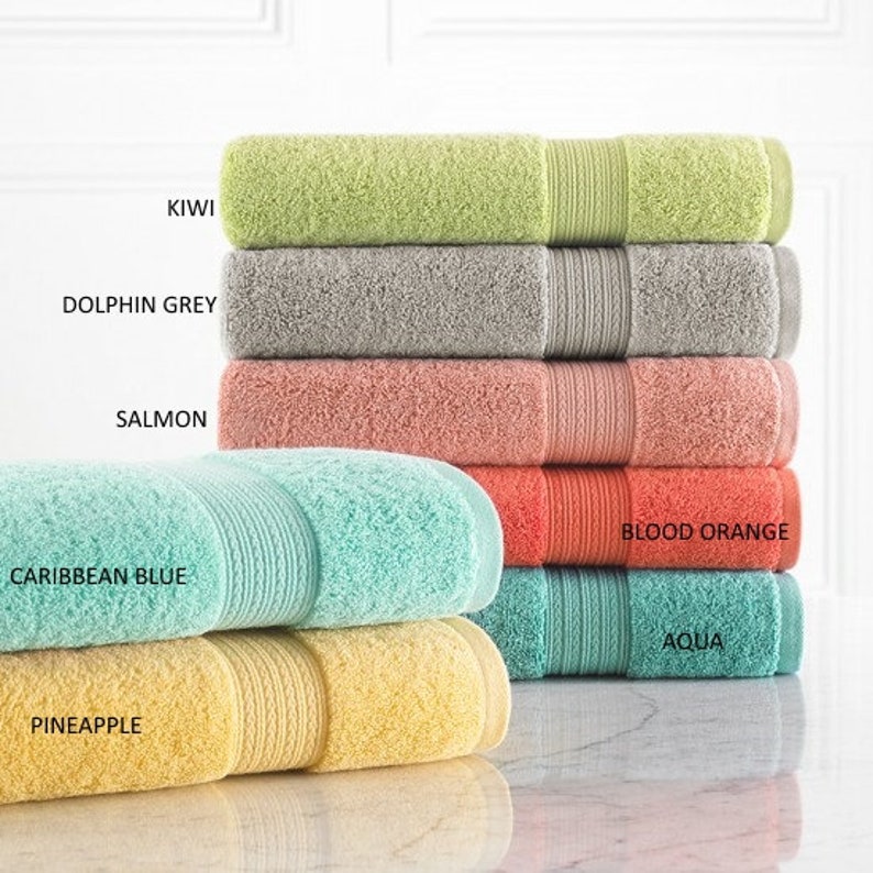 May include: A stack of six bath towels in various colors, including kiwi, dolphin grey, salmon, caribbean blue, blood orange, and aqua. The towels are folded and stacked on a white surface.