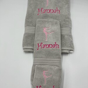 May include: A set of three gray towels with pink embroidery. The towels feature a stylized dancer and the name "Hannah".