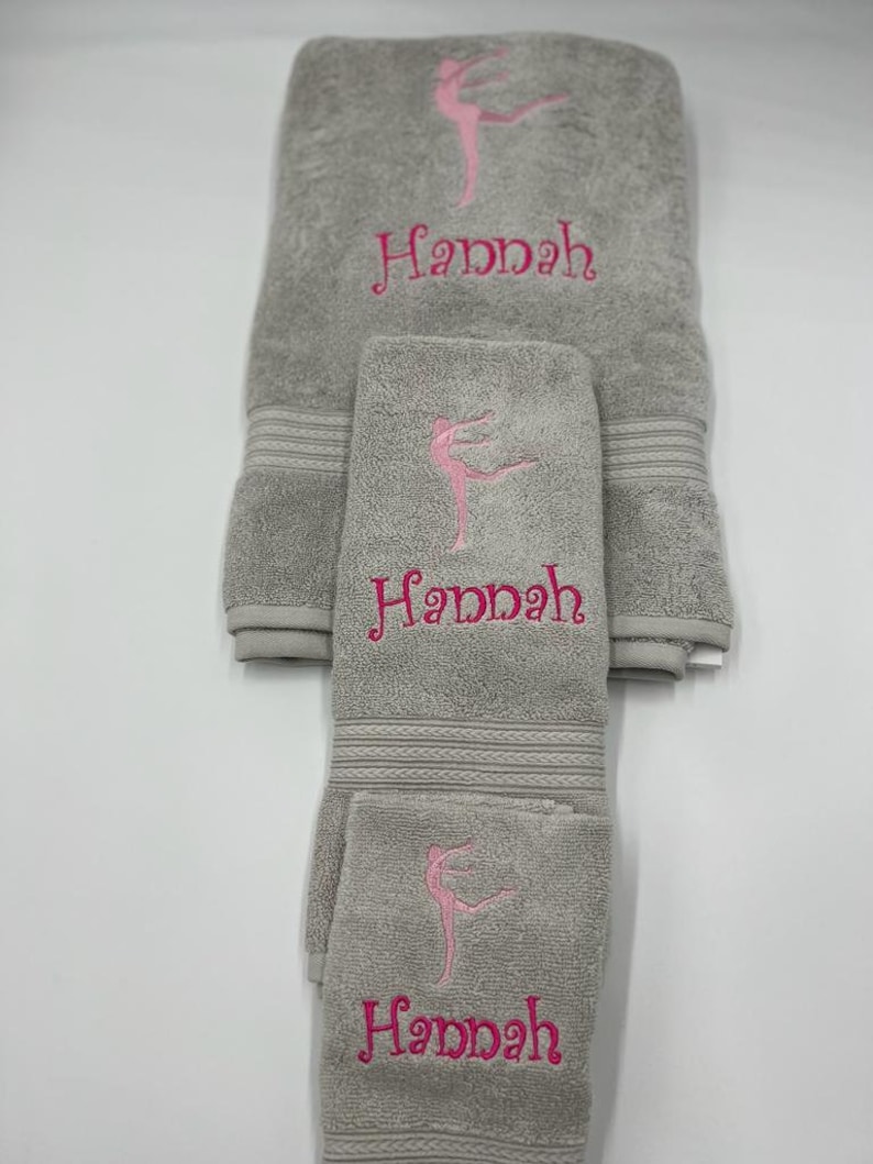 May include: A set of three gray towels with pink embroidery. The towels feature a stylized dancer and the name "Hannah".