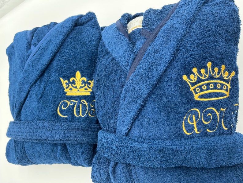 May include: Two navy blue bathrobes with gold embroidered crowns and initials. The robe on the left has the initials "CW" and the robe on the right has the initials "PR".