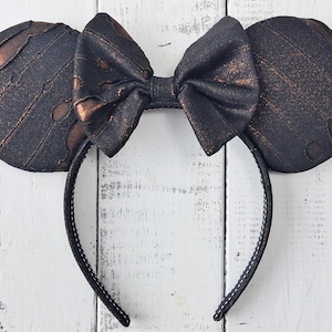 May include: Black and bronze-colored mouse ear headband with a bow. The ears have a distressed look with bronze accents. The bow is centered and matches the ear color. The headband is black with white stitching.