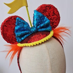 May include: A headband featuring two round ears covered in red sequins, a central bright blue sequined bow, and a yellow pom-pom trim. A yellow flag on a thin stem extends from the top, and orange feathers are attached to the sides of the band.