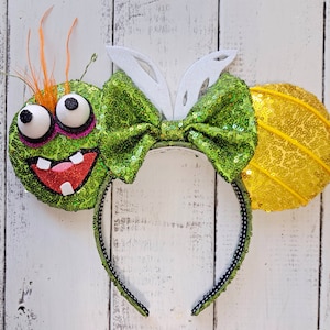 May include: A whimsical headband featuring a green sequined monster with large googly eyes, a red glittery mouth, and orange feather accents. The headband includes a green sequined bow, white bunny ears, and a yellow sequined ear.