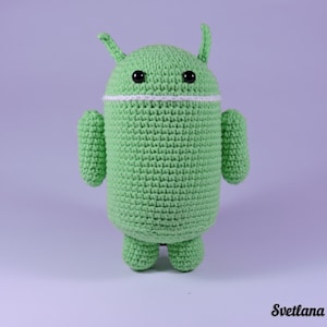 Könnte beinhalten: Grün gehäkeltes Plüschtier in Form des Android-Roboter-Maskottchens. Das Spielzeug hat schwarze Augen, einen weißen Mund und antennenartige Ohren.