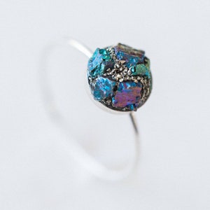 May include: A silver ring featuring a blue, green and purple iridescent gemstone. The gemstone is composed of many small, irregular pieces of crystal.