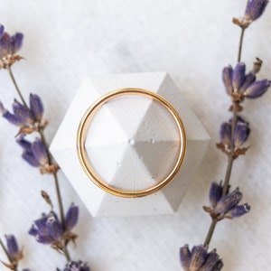 May include: A gold ring sits on a white geometric ring holder, surrounded by dried lavender sprigs on a white marble background.