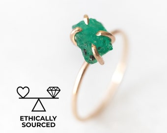 Puede incluir: Un anillo de oro con una piedra preciosa de esmeralda en bruto. La esmeralda es de color verde intenso y tiene una textura rugosa y natural. El anillo es simple y elegante, y sería un regalo único y significativo.