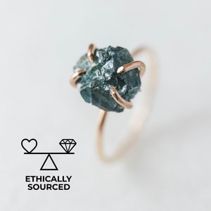 May include: A gold ring with a raw, blue-green gemstone set in the center. The ring is simple and elegant, with a delicate band.