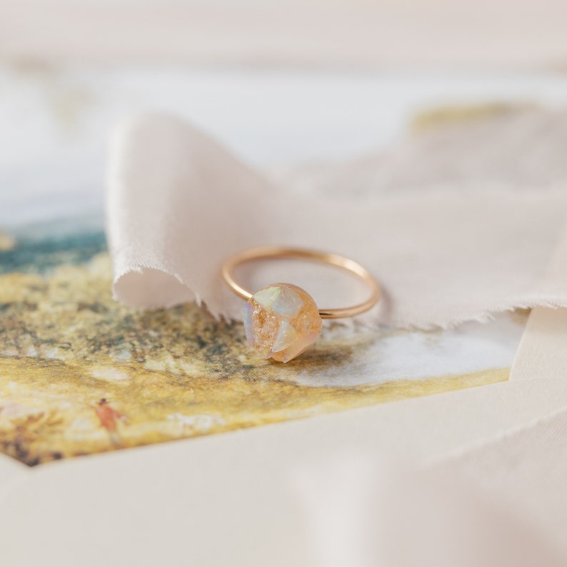 May include: A gold ring featuring a large, irregular, iridescent, light-coloured gemstone. The ring is resting on a piece of white fabric.