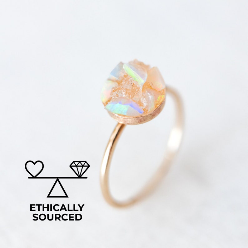 May include: A gold ring featuring a round opal gemstone set in the centre. The opal displays a mix of white, pink, and iridescent colours.