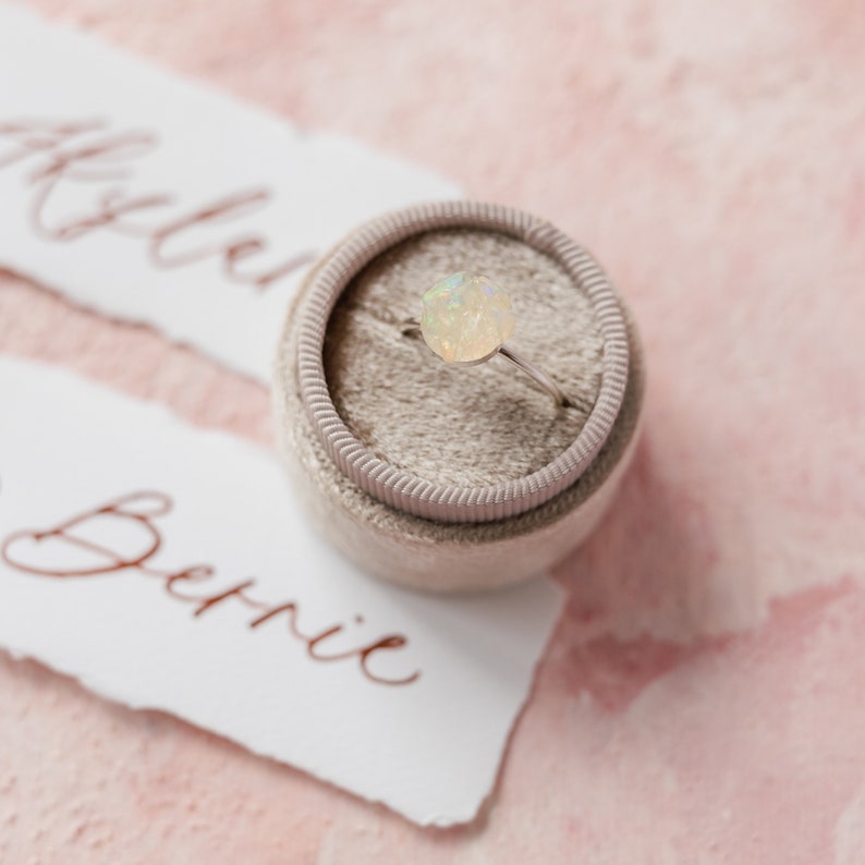 May include: A ring with a white opal gemstone set in a silver band, displayed in a small grey velvet ring box.