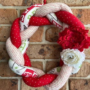 May include: A red and white Christmas wreath with a white and green fabric print that says "Merry Christmas" and has white stars. The wreath is decorated with red and white fabric flowers.