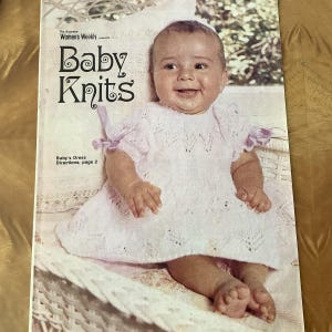 Può includere: Una copertina di rivista con un bambino che indossa un vestito bianco lavorato a maglia con un bordo rosa. Il testo della copertina recita "Baby Knits" e "Baby's Dress Directions, page 2".