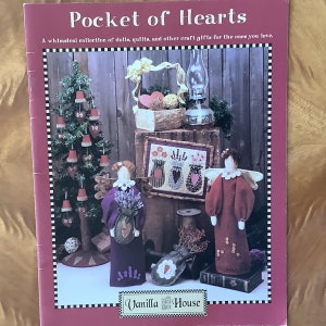 May include: A book cover with the title "Pocket of Hearts" and a whimsical illustration of handmade dolls, quilts, and other craft gifts. The cover features a red background with black and white checkered borders. The book is published by Vanilla House.