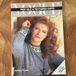 May include: Vogue Knitting Library book cover featuring a woman with red hair wearing a blue and green fair isle knit sweater. The book title is "Fair Isle & Textured Knits" and the author is Christina Probert.