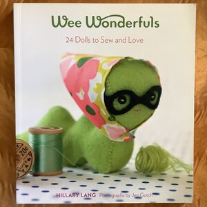 Peut inclure: Un livre intitulé "Wee Wonderfuls: 24 Dolls to Sew and Love" présentant une poupée verte et rose avec un masque noir pour les yeux. La poupée est assise sur un fond blanc à pois bleus.