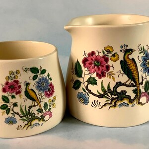 May include: A cream-coloured ceramic creamer and sugar bowl set with a floral and bird design. The design features pink, blue, yellow, and green flowers and birds on a cream background.