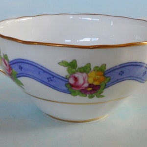 May include: A white teacup with a blue ribbon and floral design. The cup has a gold rim and handle.