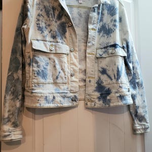 May include: A light-colored denim jacket with a blue tie-dye pattern. The jacket has two chest pockets, two side pockets, and button closures. The jacket is hanging on a black hanger.