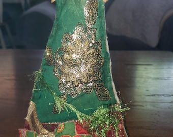 May include: A decorative Christmas tree with a rustic, vintage aesthetic. The tree features a green fabric body adorned with gold sequins and a gold star topper. A red and green patterned bow and gold trim add festive detail.