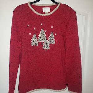 Könnte beinhalten: Ein roter Weihnachtspullover mit strukturiertem Strickdesign. Der Pullover zeigt gestickte Weihnachtsbäume und Schneeflocken. Der Halsausschnitt, die Manschetten und der Saum sind mit einer cremefarbenen, geflochtenen Bordüre versehen. Der Pullover hängt an einem schwarzen Kleiderbügel.