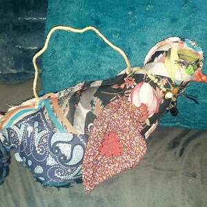 May include: A handmade fabric bird ornament. The bird is constructed from various patterned fabrics in shades of blue, red, and floral prints. It has a yellow string for hanging and a beaded necklace. The bird's beak is orange, and its eyes are green.