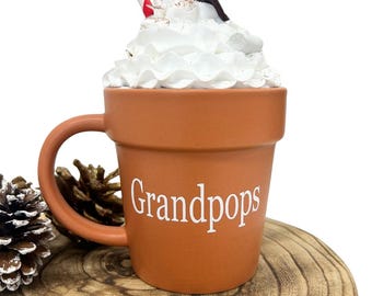May include: A brown ceramic mug shaped like a flower pot with a white handle. The mug has white lettering that says "Grandpops". The mug is filled with a white whipped cream topping and two red and white striped straws.