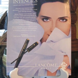 May include: A Lanc&ocirc;me mascara advertisement featuring a woman with dark hair and a white shirt. The text on the ad reads "Intencils Lash by Lash Full Intensity Mascara" and "Lash by lash...the ultimate in intensity." Two black mascara wands with gold accents are shown on the ad.