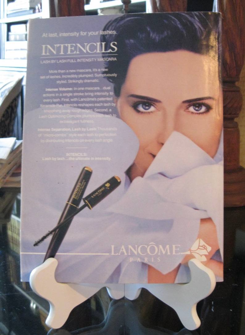 May include: A Lanc&ocirc;me mascara advertisement featuring a woman with dark hair and a white shirt. The text on the ad reads "Intencils Lash by Lash Full Intensity Mascara" and "Lash by lash...the ultimate in intensity." Two black mascara wands with gold accents are shown on the ad.