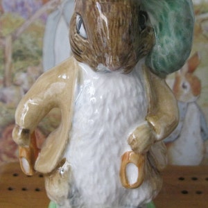 May include: A ceramic figurine of Peter Rabbit wearing a green hat and brown jacket. He is holding a brown and white object in his right hand.