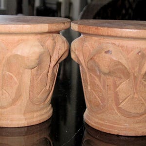 Könnte beinhalten: Zwei dekorative Holzsockel mit hellbraunem Finish. Jeder Sockel hat eine runde Oberseite und geschnitzte florale Seiten. Die Sockel sind etwa 20 cm hoch.