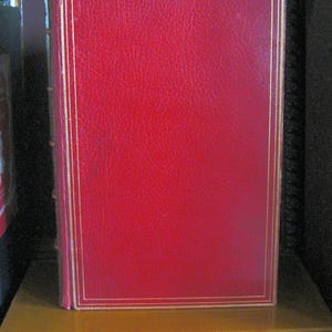 Peut inclure: Un livre relié en cuir rouge avec des bordures dorées. Le livre est posé sur une étagère avec d'autres livres. La couverture est rouge uni avec un bord doré. La tranche est visible, montrant les pages et la reliure.