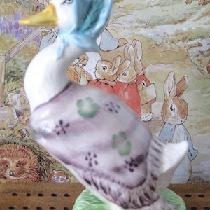 Può includere: Statuetta in ceramica di un'oca bianca con un cappello blu e uno scialle lavanda con decorazioni floreali verdi. L'oca è su una base verde con un accento giallo. Lo sfondo presenta illustrazioni di conigli e altre creature del bosco.